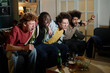 © AnnaStills - Multi-ethnic group of four young men sitting on couch in living room enjoying watching sports event on TV and supporting favorite team