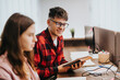 © qunica.com - Two young professionals, a man and a woman, discuss work using digital devices in a well-lit, modern office environment, exemplifying teamwork and business strategy.