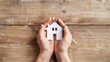 © Shahriyar - Two hands holding a paper house model on a wooden table, in the style of a real estate concept.