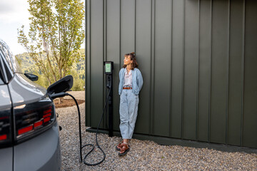 Woman waits for electric car to be charged standing near a charging station leaning to a house wall. Concept of modern lifestyle and smart technologies