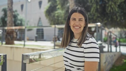 Wall Mural - A young attractive brunette woman smiles in an urban park setting on a sunny day wearing a striped shirt.