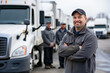 © Татьяна Евдокимова - Confident male truck driver with arms crossed in focus, with his supportive team and trucks blurred in the background