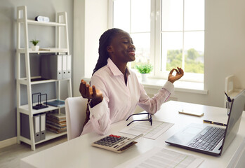 Young African American woman who works as business auditor or accountant practices stress management on workdays, sits at her office desk with laptop, does meditation, breathes, calms down and relaxes