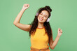 © ViDi Studio - Young woman of African American ethnicity wears yellow tank shirt top listen to music in headphones raise up hands dance isolated on plain pastel green background studio portrait. Lifestyle concept.