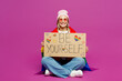© ViDi Studio - Full body young happy smiling lesbian woman wrapped in rainbow flag sits hold cardboard card with be yourself title text isolated on plain purple background. Pride day June month love LGBTQ concept.