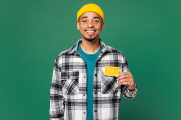Wall Mural - Young smiling fun man of African American ethnicity he wear shirt blue t-shirt yellow hat hold in hand mock up of credit bank card isolated on plain green background studio portrait Lifestyle concept