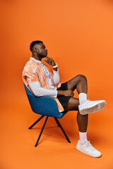 Wall Mural - Fashionable African American man sitting in a stylish chair against a bright orange background.