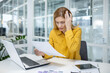 © Liubomir - Businesswoman in a yellow shirt appearing stressed while reviewing documents at her desk with a laptop and paperwork in a modern office.