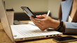 © Happy Photo - A woman is sitting at a desk with a laptop and a cell phone. She is holding a cell phone in her hand and she is typing on the laptop. The scene suggests that she is working or studying