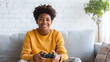 © James - Happy teenage african american boy in yellow sweatshirt sitting on sofa playing video games with controller in bright living room, Copy Space