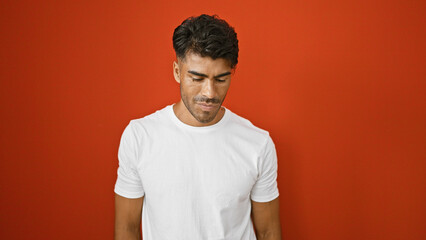 Wall Mural - A young hispanic man with a beard stands against an isolated red wall, looking pensive in casual attire.