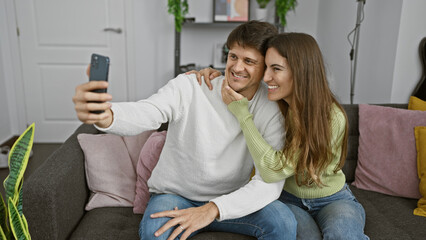 Poster - A smiling couple takes a selfie on a couch in a cozy living room, reflecting a warm, loving home atmosphere.