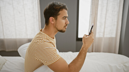 Poster - A young hispanic man with a beard thoughtfully uses his smartphone in a bedroom with curtains in the background.