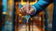 © Zoran Karapancev - A hand in a suit holds a key on a keychain in front of a blurred doorway of a modern building. Copy space, aspect ratio 16:9