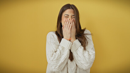 Canvas Print - A cheerful young hispanic woman in a white sweater laughing with hands on her face against a yellow background.