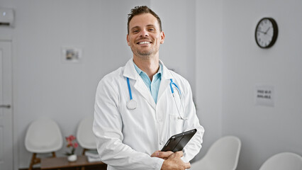 Sticker - Smiling hispanic man in white lab coat with stethoscope standing in a clinic holding a tablet.