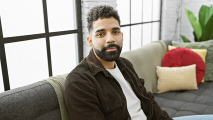 Sticker - Portrait of a relaxed young man with a beard sitting on a couch in a modern apartment living room.