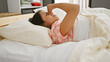 © Krakenimages.com - A young hispanic woman in a bedroom setting, looking tired as she relaxes on a bed with white bedding.