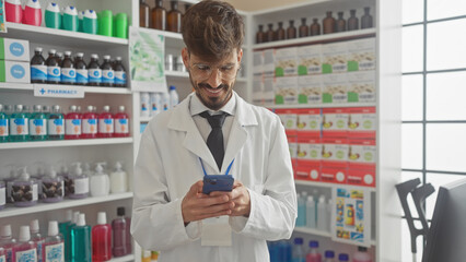 A smiling young man with a beard, wearing glasses and lab coat, using a smartphone in a brightly lit pharmacy full of healthcare products.