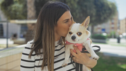 Wall Mural - A beautiful young hispanic woman holding and kissing her pet chihuahua in an urban park on a sunny day.