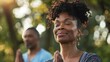 © iuricazac - A woman with curly hair wearing a blue top has her eyes closed and is smiling while a man in a blue shirt stands behind her with his hands together in a prayer position both in a peaceful outdoor