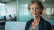 © iuricazac - A poised middle-aged woman with short gray hair wearing a dark blazer and a light-colored shirt sitting in a modern office with a whiteboard in the background.