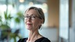© iuricazac - A woman with short blonde hair wearing glasses and a black top standing in a modern office with blurred greenery in the background.