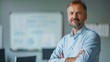 © Z A C - A man with a beard and graying hair wearing a light blue shirt standing confidently with his arms crossed in front of a whiteboard with writing on it in a professional office setting.