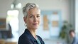 © iuricazac - Woman with short gray hair wearing a dark blazer smiling at the camera standing in a brightly lit room with a blurred background.