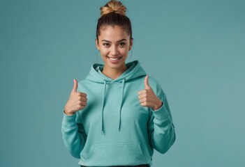 Wall Mural - A smiling young woman in a turquoise hoodie gives two thumbs up, signaling positivity and a relaxed demeanor.