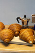 © brajianni - Vertical photo. Delicious warm tender croissant on wooden board on table. Coffee maker near gray kitchen wall. Concept, lunch, dinner, food, eating. Bakery with fresh pastries. Copy space. French cafe