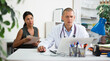 © JackF - Focused doctor sitting at table with laptop, consulting female patient and writing prescription at clinic..
