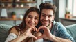 © inthasone - A young couple making a heart sign with their hands at home, sitting on the sofa in the living room, and smiling at the camera. A concept of love. Celebrating Valentine's Day
