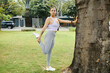 © DragonImages - Woman in athletic wear stretching her leg while leaning against a tree in a sunny park