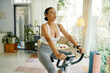 © DragonImages - A woman in workout attire exercising on a stationary bike in a well-lit living room with fitness equipment