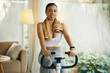 © DragonImages - A woman in activewear resting on an exercise bike with a towel around her neck in a cozy living room