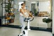 © DragonImages - A woman in workout clothes using a stationary bike in a bright, modern living room with plants and a cozy atmosphere