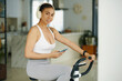 © DragonImages - A woman in workout attire using her phone while exercising on a stationary bike, wearing headphones in a stylish interior