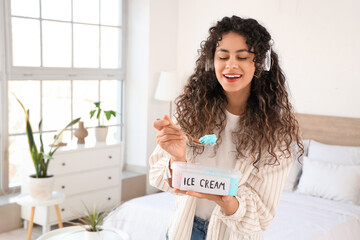 Wall Mural - Beautiful young African-American woman in headphones with basket of ice cream in bedroom