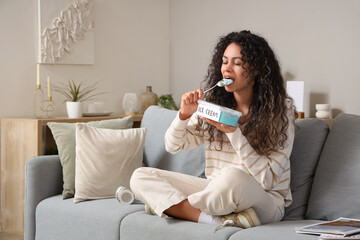 Sticker - Beautiful young African-American woman eating ice cream and sitting on sofa in living room