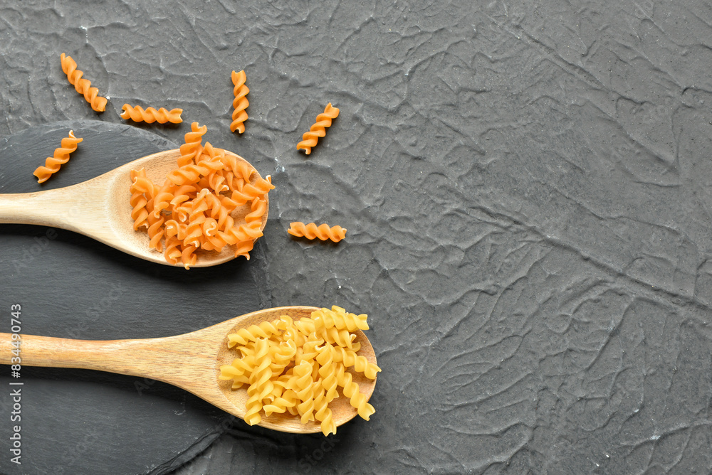Wooden spoons with different tasty uncooked fusilli pasta on dark background