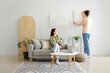 © Pixel-Shot - Young man hanging painting on light wall and his wife at home