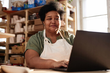 Wall Mural - Warm toned portrait of African American senior woman using laptop in art studio lit by sunlight copy space