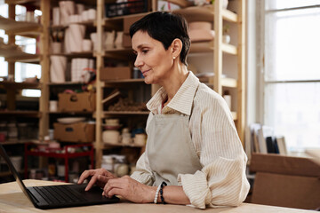 Wall Mural - Side view portrait of elegant senior woman using computer in cozy art studio and looking up creative ideas copy space