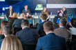 © Anton Gvozdikov - Attendees at a business conference listening to a panel discussion with speakers on stage, engaging in professional networking.