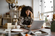 © sofiko14 - Young curly woman potter taking order for purchase of tableware using phone and laptop. Happy female with new purchase in ceramics shop sitting at table against background of shelves with dishes
