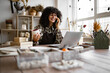 © sofiko14 - Happy female with new purchase in ceramics shop sitting at table against background of shelves with dishes. Young curly woman potter taking order for purchase of tableware using phone and laptop