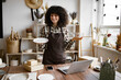 © sofiko14 - Attractive curly woman wearing brown apron receiving goods at modern decor store. Professional seller checking for no damages on cup and plate that holding in hands.