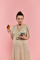 A young pregnant woman in a dress holding a cell phone and a credit card against a pink background.