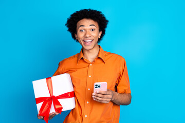 Poster - Photo of attractive funny guy wear orange shirt rising gift box texting gadget emtpy space isolated blue color background